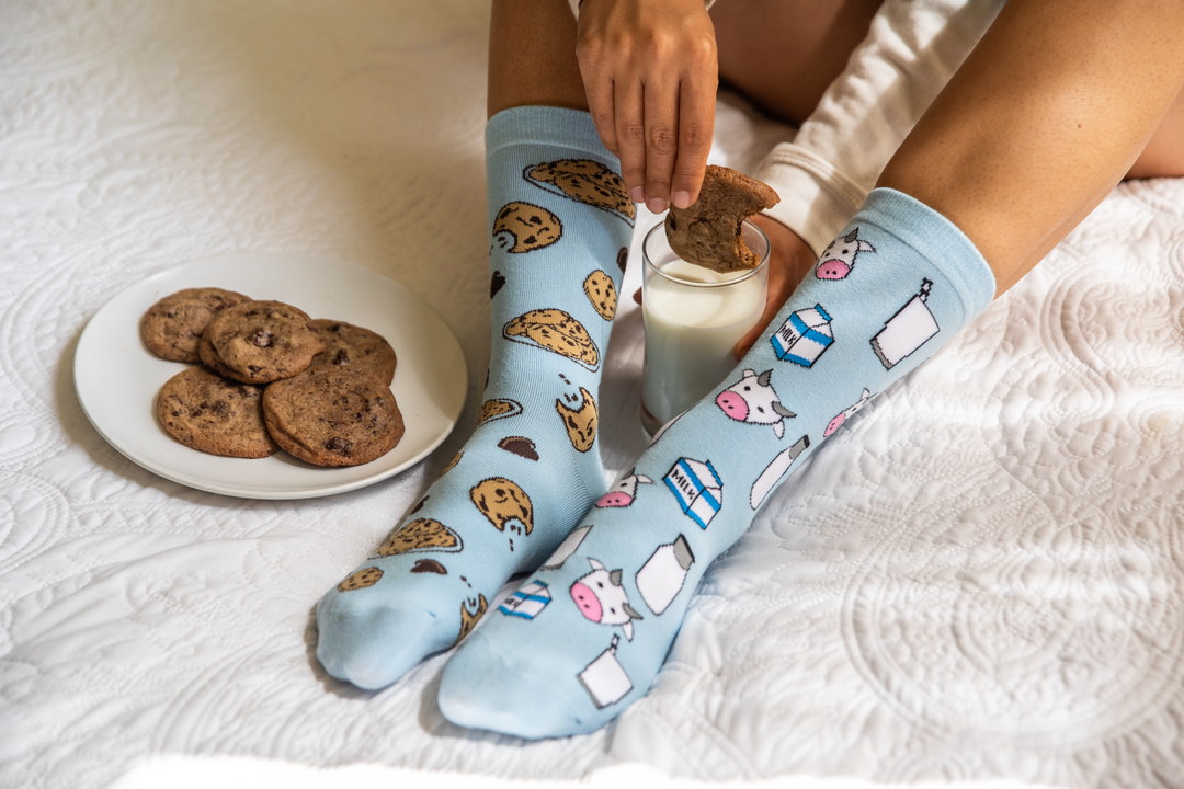 Person wearing a cookie printed sock and a milk and cow printed sock while dipping a bitten cookie into a glass of milk