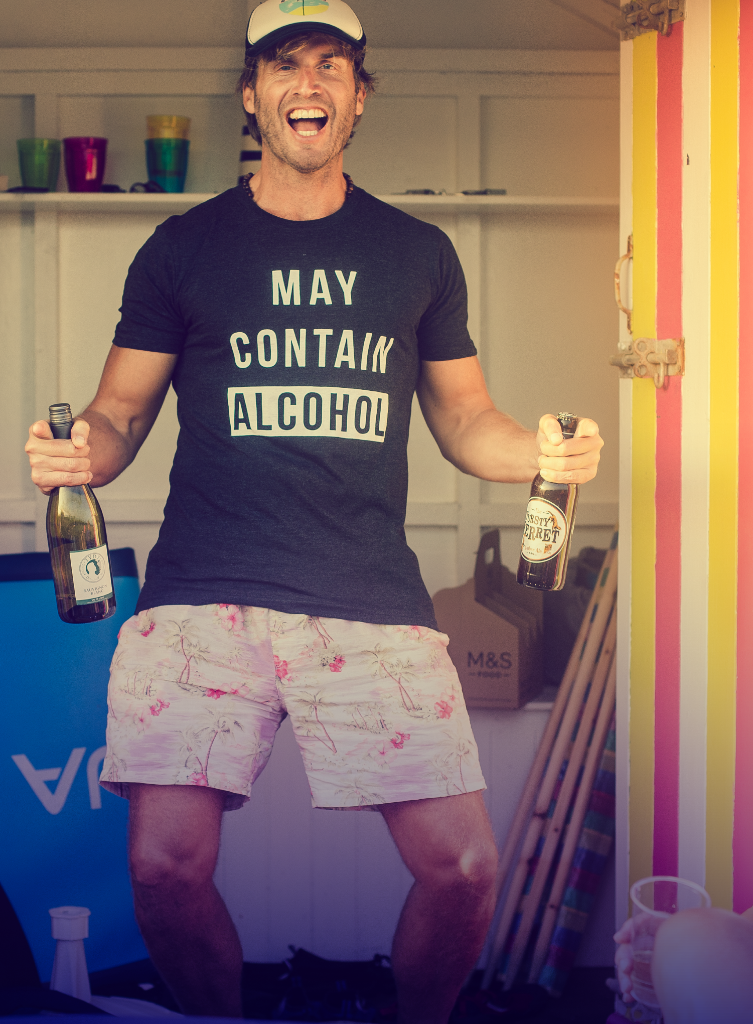 Man with a hat, olding a beer bottle and a champagne bottle, wearing a may contain alcohol, printed shirt