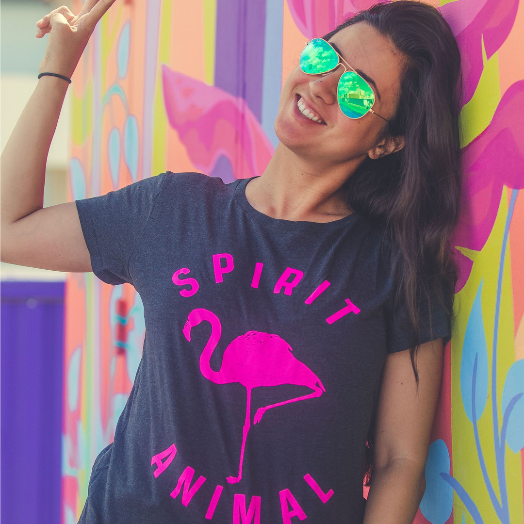 Woman wearing sunglasses doing the peace sign, wearing a dark grey shirt with a pink print out that says spirit animal around the picture of a flamengo