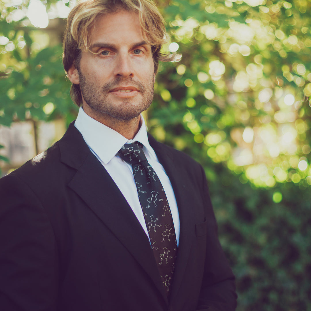 Man in a suit with a patterned tie standing outdoors with greenery in the background